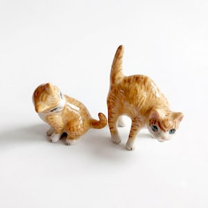 May include: Two small ceramic cat figurines with a glossy finish. One cat is sitting in a curled position, while the other stands with its back arched. Both figurines feature orange tabby stripes, white accents on the paws and face, and blue eyes.