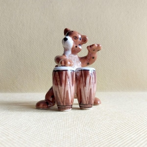 Puede incluir: Figura de cerámica de un oso tocando los tambores conga. El oso es marrón con manchas negras y detalles blancos. Los tambores son marrones con un borde blanco y un patrón vertical.