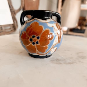 May include: A small ceramic vase with two handles, featuring a black rim and base. The body is decorated with a large orange flower with a blue center, set against a light blue background with white vine-like patterns wrapping around the sides.