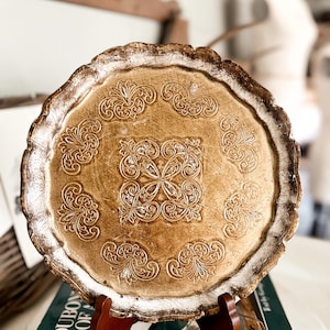 May include: A round decorative plate with a scalloped edge, featuring a metallic gold finish and a central square embossed pattern surrounded by eight smaller symmetrical floral-like motifs, all highlighted with silver-toned distressed accents.