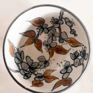 May include: A round ceramic bowl with a cream-colored base featuring a hand-painted botanical design. The pattern consists of dark gray branches with light gray blossoms and brown leaves, finished with a dark gray rim around the top edge.