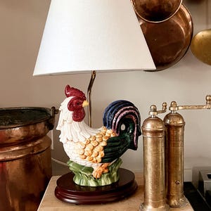 May include: A table lamp featuring a ceramic rooster base with a white, red, and green plumage, standing on a dark wood oval pedestal. The lamp has a simple white conical fabric shade and a thin metallic neck extending from the rooster's back.