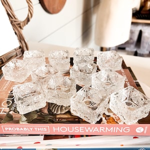 May include: A collection of twelve clear, square glass salt cellars featuring a pressed geometric pattern on the exterior. Each small, open-top vessel has thick walls and a textured base, arranged in a grid formation on top of stacked books.