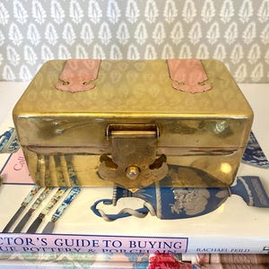 May include: A rectangular brass box with a hinged lid and a clasp closure. The box has pink accents on the lid and a decorative latch. The box is sitting on top of a book titled "Collector's Guide to Buying Blue Pottery & Porcelain".