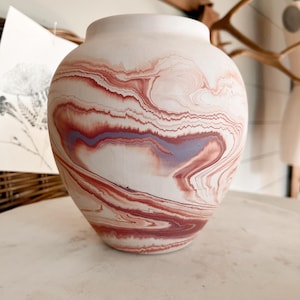May include: A rounded ceramic vase featuring a matte finish with a marbled pattern of swirling terracotta, dusty rose, and muted lavender tones. The vessel has a wide, bulbous body that tapers toward a smooth, circular opening at the top.