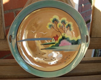 Puede incluir: Un plato de servir de cerámica vintage con una escena pintada a mano de un lago, una casa y árboles. El plato tiene un borde verde y un fondo dorado.