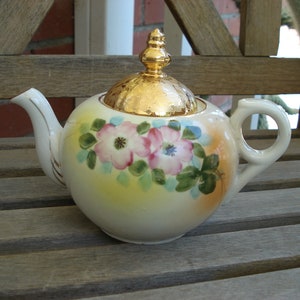 May include: A white porcelain teapot with a gold lid and floral design. The teapot has a round body and a curved spout. The flowers are pink and white with green leaves.
