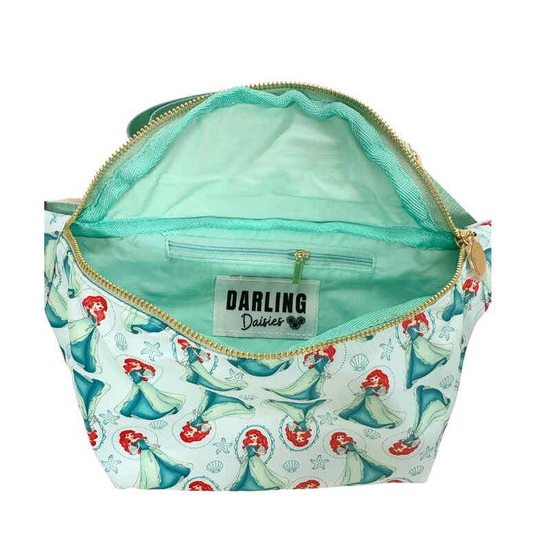 Puede incluir: Bolso de mano azul claro abierto con cremallera dorada e interior verde menta. El exterior presenta un patr&oacute;n repetido de una sirena pelirroja con un vestido azul, conchas marinas y estrellas de mar. La etiqueta interior dice "DARLING Daisies".
