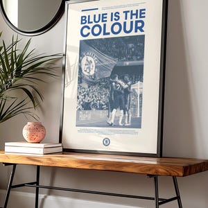 May include: A framed poster with a blue and white color scheme. The poster features a black and white photo of a crowd of people at a sporting event. The text "BLUE IS THE COLOUR" is printed in bold, blue letters at the top of the poster.