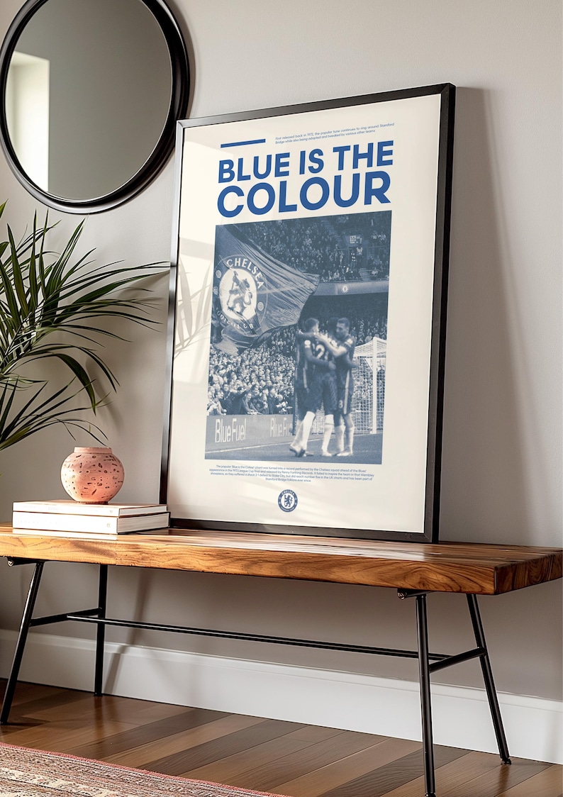 May include: A framed poster with a blue and white color scheme. The poster features a black and white photo of a crowd of people at a sporting event. The text "BLUE IS THE COLOUR" is printed in bold, blue letters at the top of the poster.