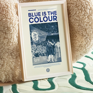 May include: A framed poster with a blue and white color scheme. The poster features a photo of a crowd of people at a soccer game, with a large flag that says "Chelsea Football Club" in the foreground. The text "Blue is the Colour" is printed in large, bold letters at the top of the poster.