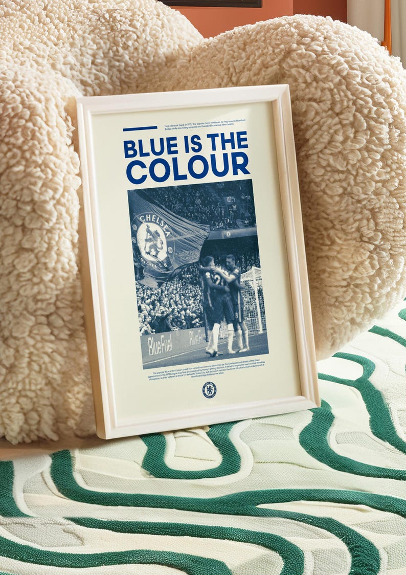 May include: A framed poster with a blue and white color scheme. The poster features a photo of a crowd of people at a soccer game, with a large flag that says "Chelsea Football Club" in the foreground. The text "Blue is the Colour" is printed in large, bold letters at the top of the poster.