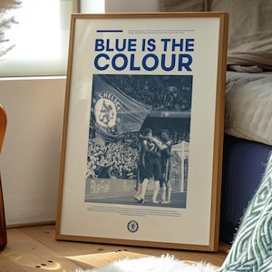 May include: A framed poster with a blue and white color scheme. The poster features a photo of a soccer game with a large blue flag that says "Chelsea" in white letters. The text "Blue is the color" is printed in bold blue letters at the top of the poster.