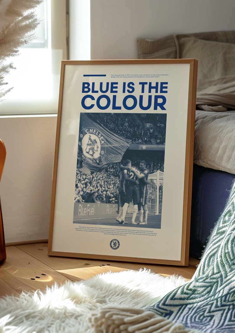 May include: A framed poster with a blue and white color scheme. The poster features a photo of a soccer game with a large blue flag that says "Chelsea" in white letters. The text "Blue is the color" is printed in bold blue letters at the top of the poster.