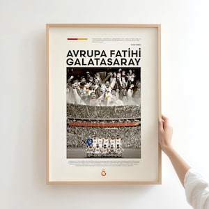 May include: Framed poster with the text "AVRUPA FATIHI GALATASARAY" in bold black letters. The poster features a sepia-toned image of a soccer team celebrating a victory, with a crowd in the background. The poster is held by a person.