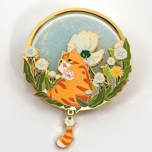 May include: A round, gold-toned pin featuring an orange tabby cat lounging among white flowers and green foliage. The background is a light blue with glitter. A dangling charm with a striped tail hangs below.