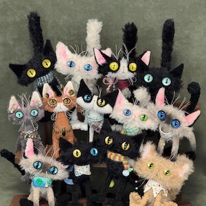 Könnte beinhalten: Eine Sammlung handgefertigter Katzenplüschtiere in verschiedenen Farben und Mustern. Jede Katze hat einzigartige Merkmale, darunter unterschiedliche Augenfarben, Accessoires wie Schals und Halsbänder sowie unterschiedliche Fellstrukturen. Sie sind auf einem Holzregal angeordnet.