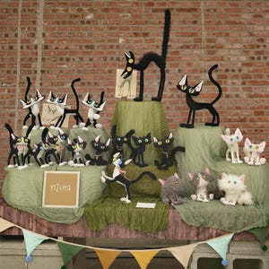 May include: A collection of handcrafted felt cat figurines in various poses, colors, and sizes, featuring large expressive eyes and long tails. A square sign with the text YIZCHO is displayed among the figures, which are arranged on green fabric.