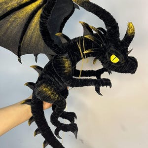 May include: A black plush dragon with gold accents on the wings, horns, and claws. The dragon has a long tail and a yellow eye. A gold chain is draped around the dragon's neck. The dragon is a fantasy-themed item.
