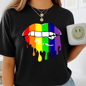 May include: Black t-shirt featuring a colorful, rainbow-themed lip design with dripping paint effect. A white mug with a smiling face is held in the hand. The person is wearing a gold necklace and blue shorts.