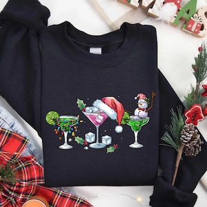May include: A black sweatshirt featuring a festive cocktail design. The graphic includes three margarita glasses, each decorated with Christmas themes: lights, a Santa hat, and a snowman. The design is set against a backdrop of holiday decorations.