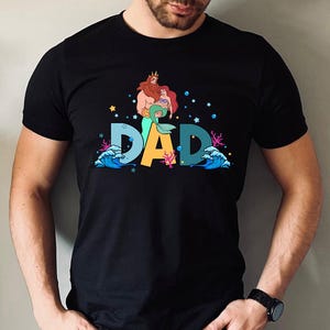 May include: Black t-shirt with a graphic of Ariel and King Triton from The Little Mermaid. The graphic says "DAD" with waves and sea life surrounding the letters.