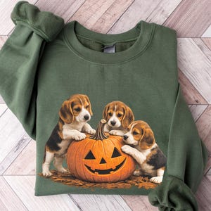 May include: An olive green crewneck sweatshirt featuring a graphic print of three beagle puppies interacting with a carved orange jack-o'-lantern pumpkin. The puppies are positioned around the pumpkin, which sits on a bed of brown autumn leaves.