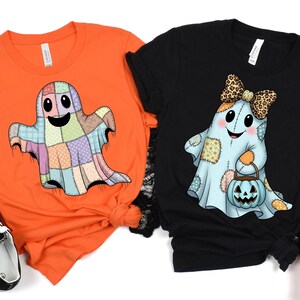 May include: Two Halloween-themed t-shirts. One is orange with a patchwork ghost graphic. The other is black with a ghost wearing a bow and holding a pumpkin bucket. Black and orange sneakers are on either side of the shirts.