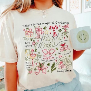 May include: A cream-coloured t-shirt with a Christmas graphic. The design features the text "Believe in the magic of Christmas" and illustrations of gifts, candy canes, a Christmas tree, and a gingerbread house. A white mug with a smiley face is also visible.
