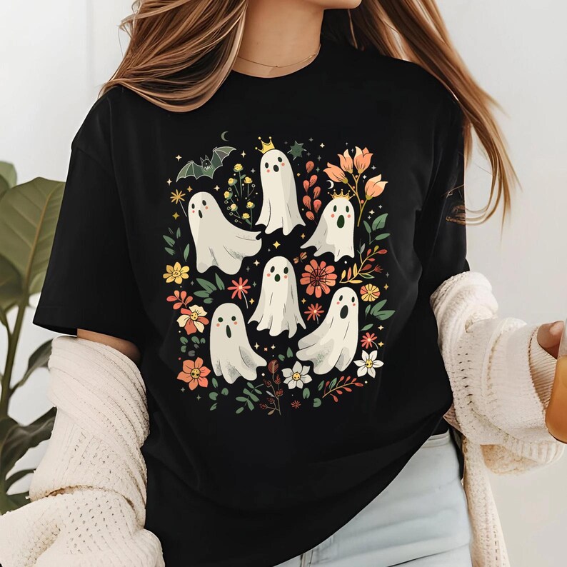 May include: Black t-shirt featuring a whimsical design of white ghosts with crowns and floral accents. The ghosts are surrounded by colorful flowers and foliage, creating a playful and festive Halloween theme. The design is centered on the shirt.