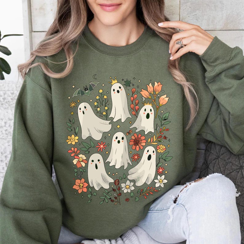 May include: Olive green sweatshirt featuring a whimsical design of cartoon ghosts with floral accents. The ghosts have various expressions and some wear crowns. A bat and crescent moon are also included in the design. The sweatshirt is a comfortable, casual style.