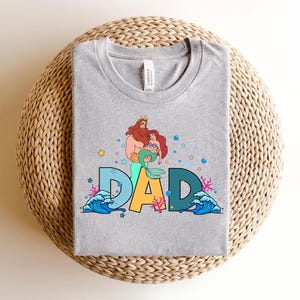May include: Gray t-shirt with a graphic of King Triton and Ariel from The Little Mermaid. The graphic includes the word "DAD" with ocean waves and sea life.