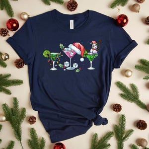 May include: Navy blue t-shirt featuring three festive cocktail designs. One is decorated with lights and a lime, another with a Santa hat, and the last with a snowman. The shirt is surrounded by Christmas decorations.