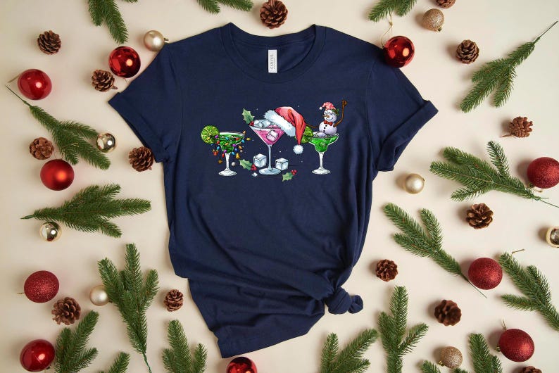 May include: Navy blue t-shirt featuring three festive cocktail designs. One is decorated with lights and a lime, another with a Santa hat, and the last with a snowman. The shirt is surrounded by Christmas decorations.