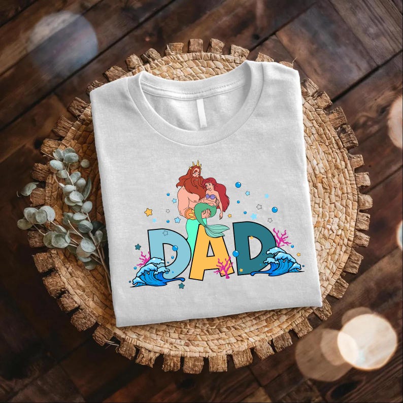 May include: White t-shirt with a graphic of a cartoon merman and mermaid with the word "DAD" in colorful letters surrounded by sea-themed imagery.