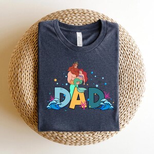 May include: Dark blue t-shirt with a graphic of a cartoon merman and mermaid with the word "DAD" in colorful letters. The graphic is surrounded by sea-themed elements like waves and coral.