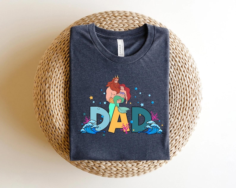 May include: Dark blue t-shirt with a graphic of a cartoon merman and mermaid with the word "DAD" in colorful letters. The graphic is surrounded by sea-themed elements like waves and coral.
