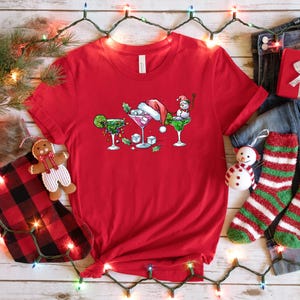 May include: A red t-shirt featuring a festive design of three cocktails, each adorned with holiday elements like a Santa hat and a snowman. The shirt is surrounded by Christmas decorations, including a stocking, gift, and string lights.