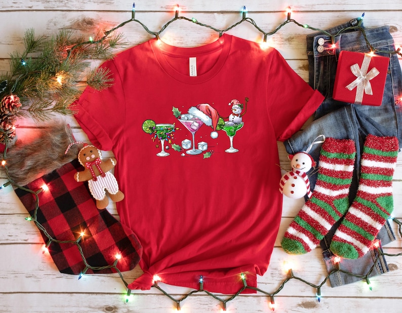 May include: A red t-shirt featuring a festive design of three cocktails, each adorned with holiday elements like a Santa hat and a snowman. The shirt is surrounded by Christmas decorations, including a stocking, gift, and string lights.