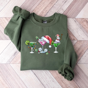 May include: An olive green sweatshirt featuring a festive cocktail design. The graphic includes three margarita glasses, each decorated with holiday elements like a Santa hat, lights, and a snowman. The design is centered on the front of the sweatshirt.