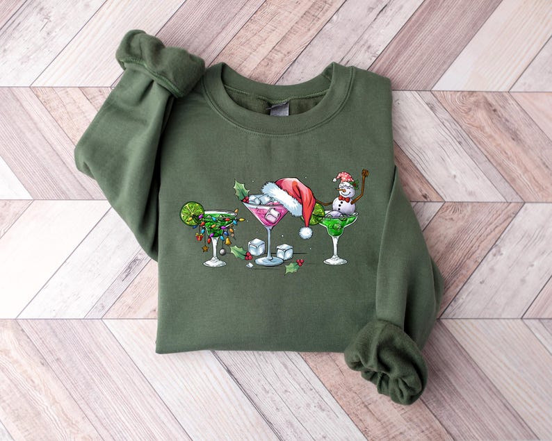May include: An olive green sweatshirt featuring a festive cocktail design. The graphic includes three margarita glasses, each decorated with holiday elements like a Santa hat, lights, and a snowman. The design is centered on the front of the sweatshirt.