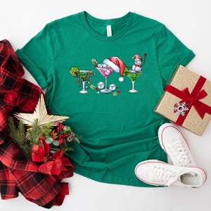 May include: A green t-shirt featuring three festive cocktail glasses with holiday decorations. One glass has a lime, another a Santa hat, and the third a snowman. A gift box, plaid scarf, star, and white sneakers complete the holiday theme.