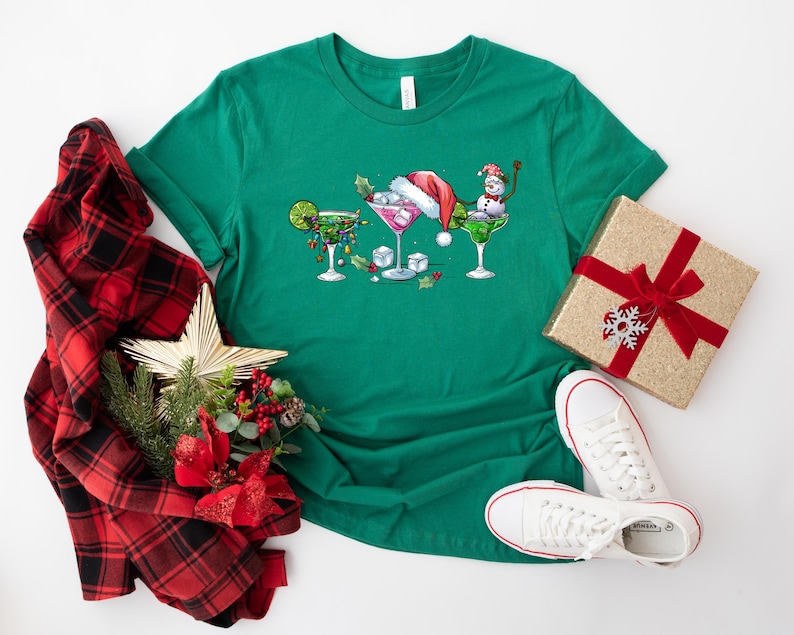 May include: A green t-shirt featuring three festive cocktail glasses with holiday decorations. One glass has a lime, another a Santa hat, and the third a snowman. A gift box, plaid scarf, star, and white sneakers complete the holiday theme.