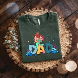 May include: A dark green t-shirt with a graphic print of a cartoon version of King Triton and Ariel from the Disney movie The Little Mermaid. The graphic includes the word "DAD" in colorful letters with bubbles and seaweed surrounding the letters.