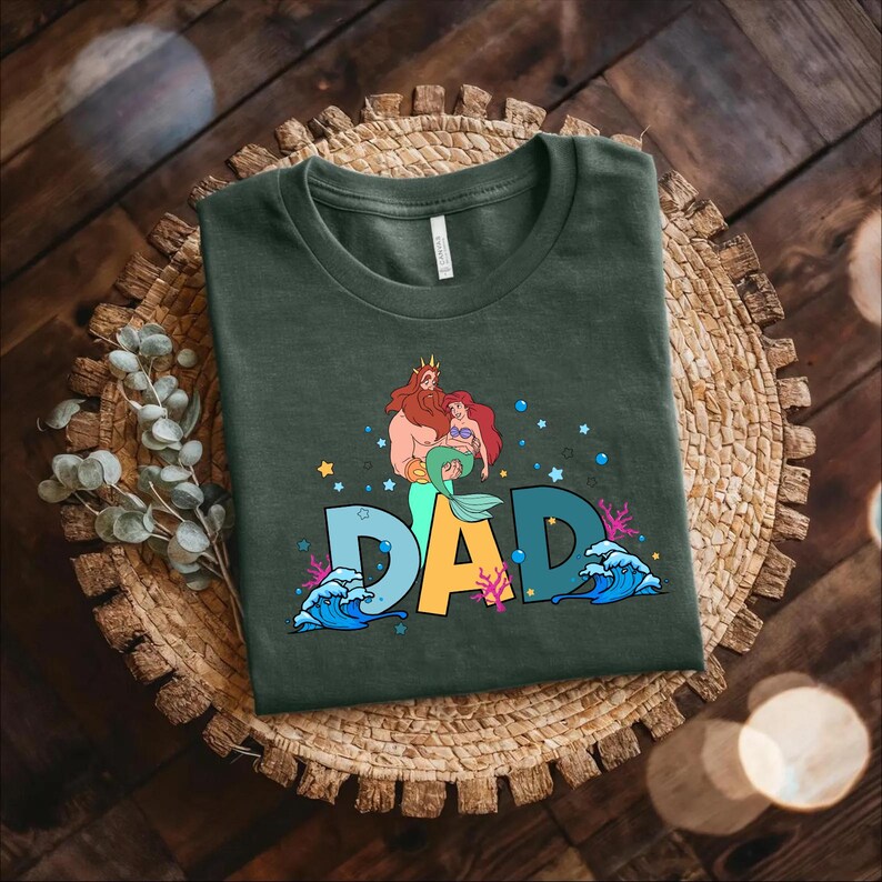 May include: A dark green t-shirt with a graphic print of a cartoon version of King Triton and Ariel from the Disney movie The Little Mermaid. The graphic includes the word "DAD" in colorful letters with bubbles and seaweed surrounding the letters.