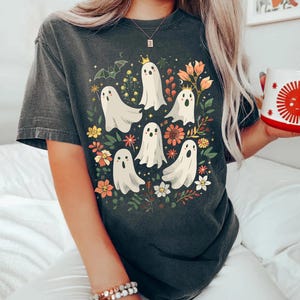 May include: Dark gray t-shirt featuring a whimsical design of white ghost figures with floral accents. The ghosts have crowns and are surrounded by colorful flowers and foliage, creating a playful and festive look.