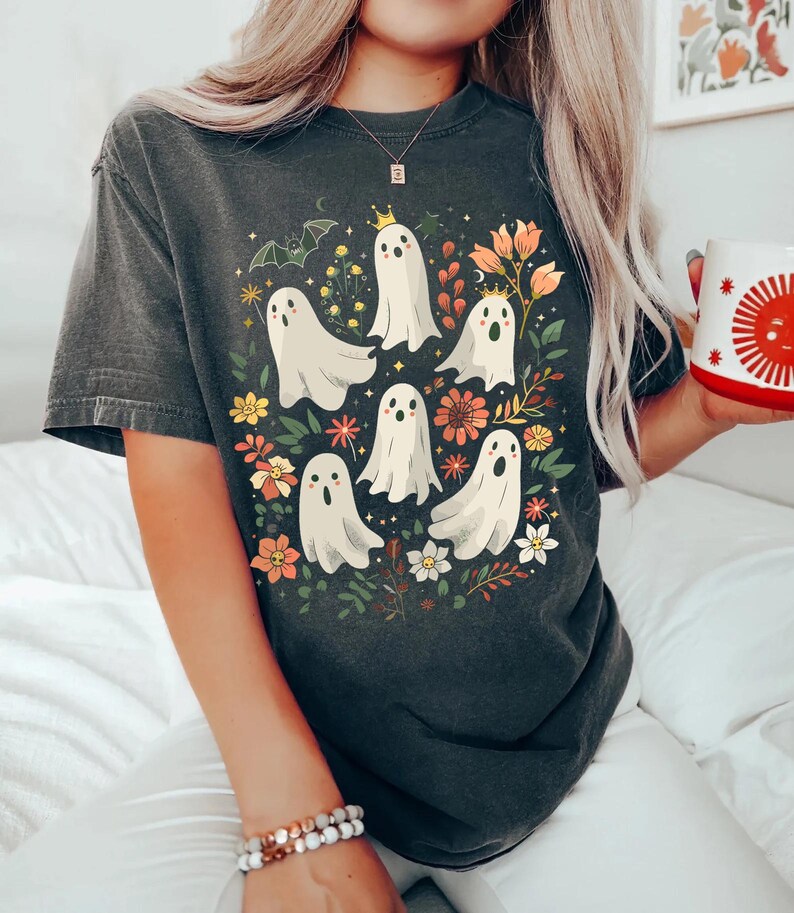 May include: Dark gray t-shirt featuring a whimsical design of white ghost figures with floral accents. The ghosts have crowns and are surrounded by colorful flowers and foliage, creating a playful and festive look.