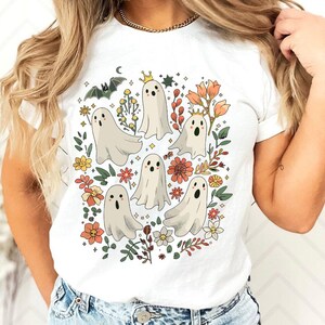 May include: White t-shirt featuring a whimsical design of cartoon ghosts surrounded by flowers, leaves, and a bat. The ghosts have crowns and various expressions. The design incorporates orange, yellow, and green floral elements.