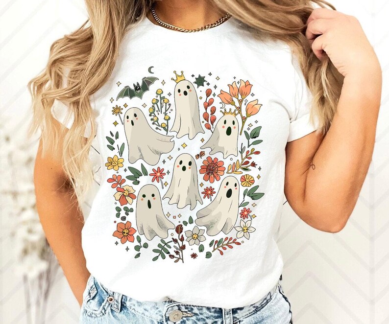 May include: White t-shirt featuring a whimsical design of cartoon ghosts surrounded by flowers, leaves, and a bat. The ghosts have crowns and various expressions. The design incorporates orange, yellow, and green floral elements.