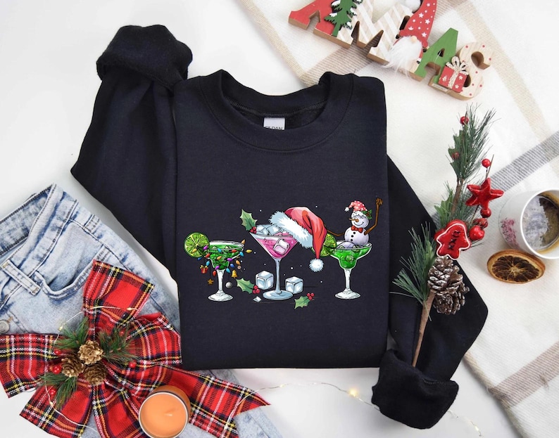 May include: A black sweatshirt featuring a festive cocktail design. The graphic includes three margarita glasses, each decorated with Christmas themes: lights, a Santa hat, and a snowman. The design is set against a backdrop of holiday decorations.